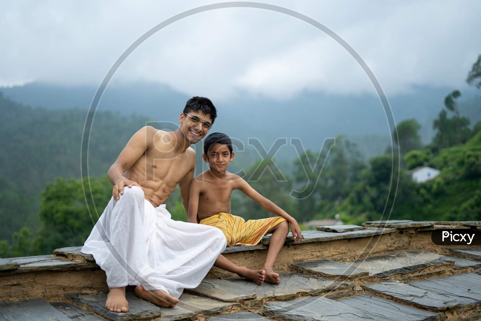 A Young Man Sitting On The Roof With His Younger Brother,Both Wearing Dhoti, Smiling Into The Camera