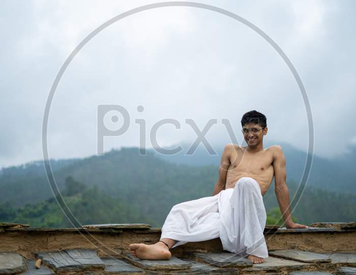 Young Shredded Man Sitting Wearing A Dhoti And Glasses With Blurred Mountains In The Background.
