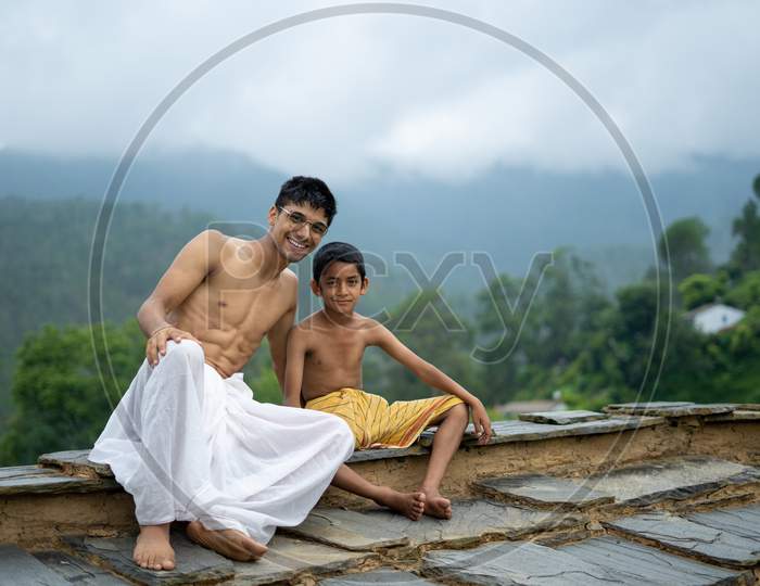 A Young Man Sitting On The Roof With His Younger Brother,Both Wearing Dhoti, Smiling Into The Camera