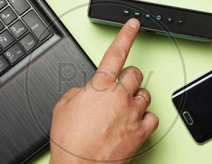 Top View Of Man'S Hand Turning On A Loudspeaker Next To Phone And Laptop. Concept Listen To Music Phone Or Computer. Flat Lay