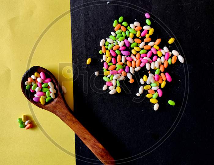 Candies In Wooden Spoon And Scattered Colored Candies Isolated On Yellow And Black Background.