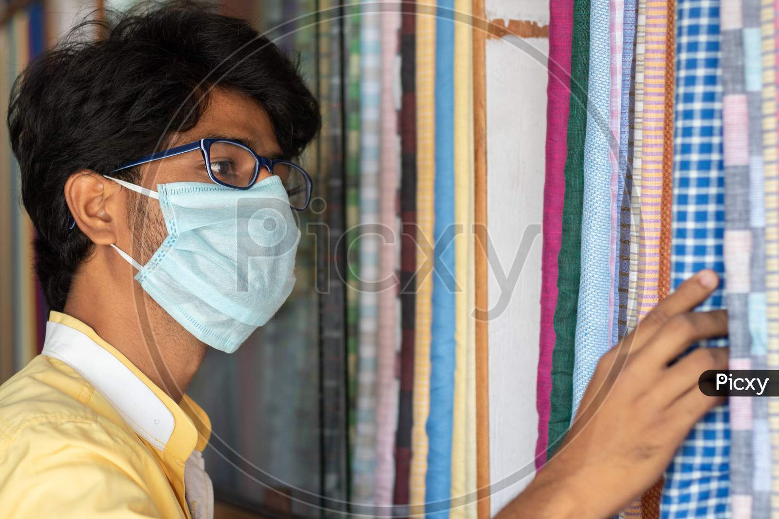 Young Man With Medical Mask Selecting Or Buying Cloth At Store During Coronavirus Or Covid-19 Pandemic - Concept Of New Normal, Business Reopen And Support Local Community After Lock Down.
