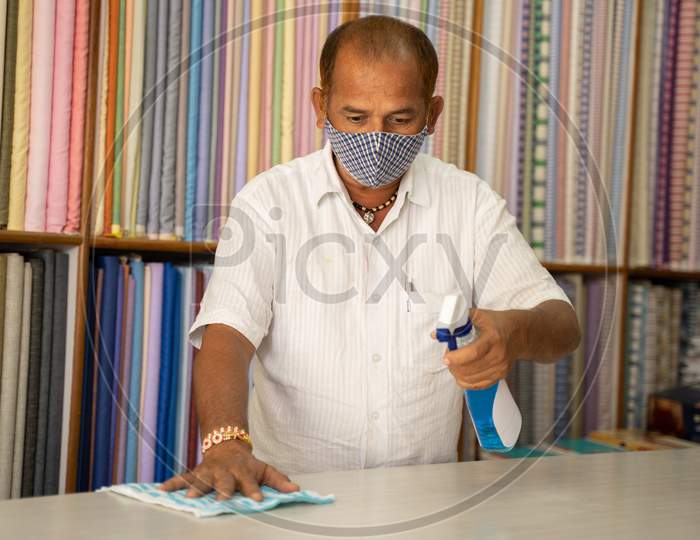 Shopkeeper In Medical Mask Disinfecting Table By Using Sanitizer - Cleaning Dust On Desk Surface With Cloth Or Disinfectant Spray, To Protect From Coronavirus Or Covid-19 Infection.