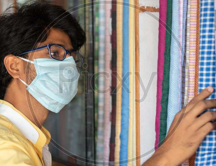 Young Man With Medical Mask Selecting Or Buying Cloth At Store During Coronavirus Or Covid-19 Pandemic - Concept Of New Normal, Business Reopen And Support Local Community After Lock Down.