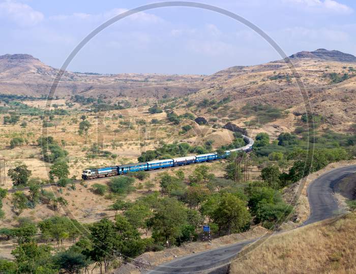 Train On A Curved Track With Background Of Hills And Sky And A Foreground Road.