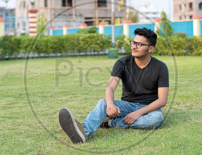 Fashion Portrait Of Indian Guy Wearing A Black T Shirt, Sits On The Ground And Poses Against The Background Of Nature. Indian Lifestyle And Fashion