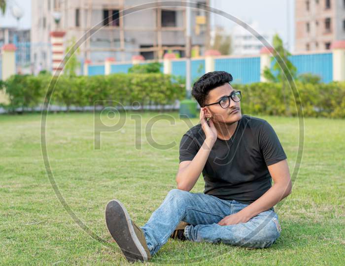 Fashion Portrait Of Indian Guy Wearing A Black T Shirt, Sits On The Ground And Poses Against The Background Of Nature. Indian Lifestyle And Fashion