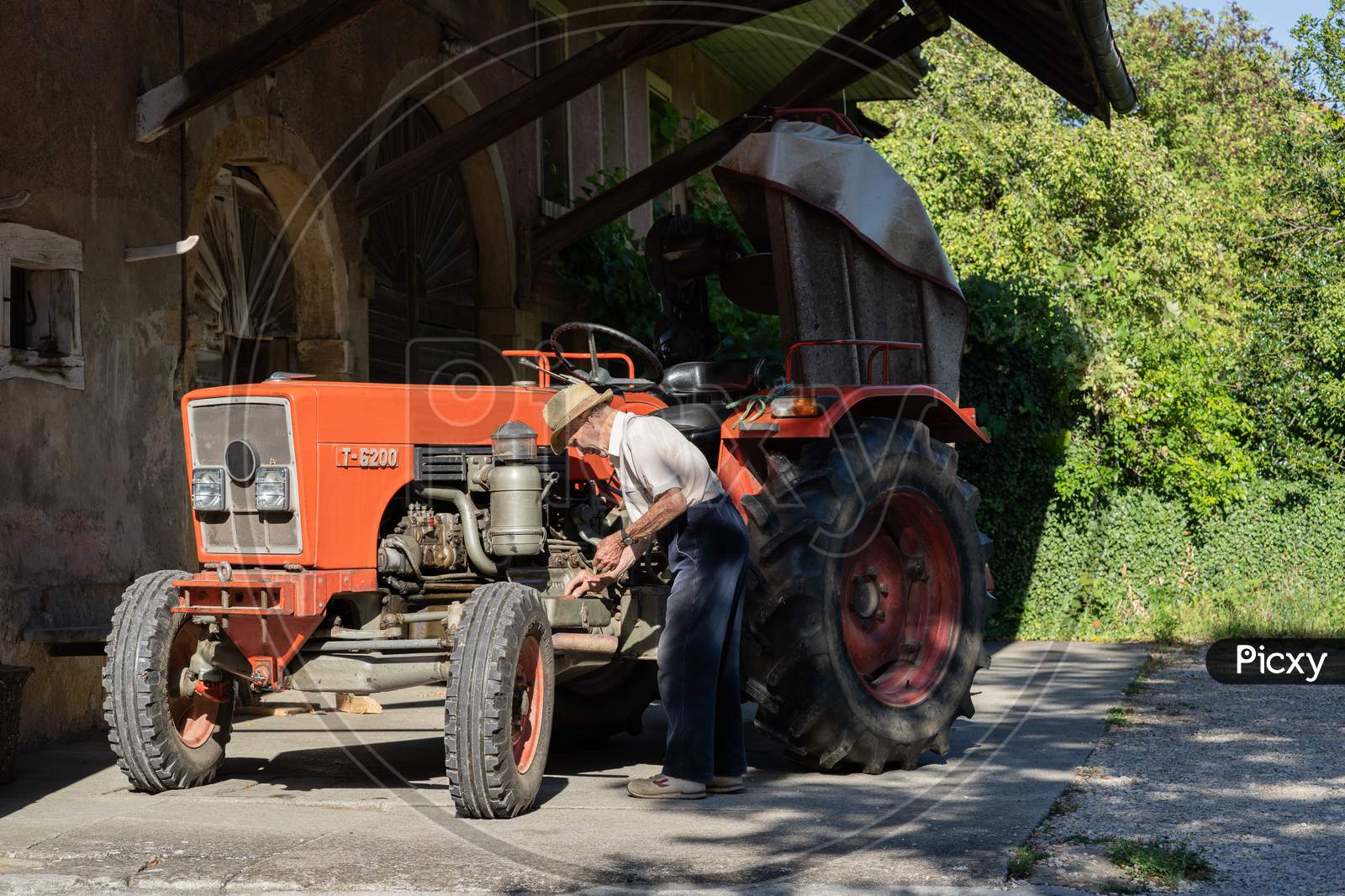 Old Farmer Working On Red Tractor Under Roof Of Farmer House In Sunshine.