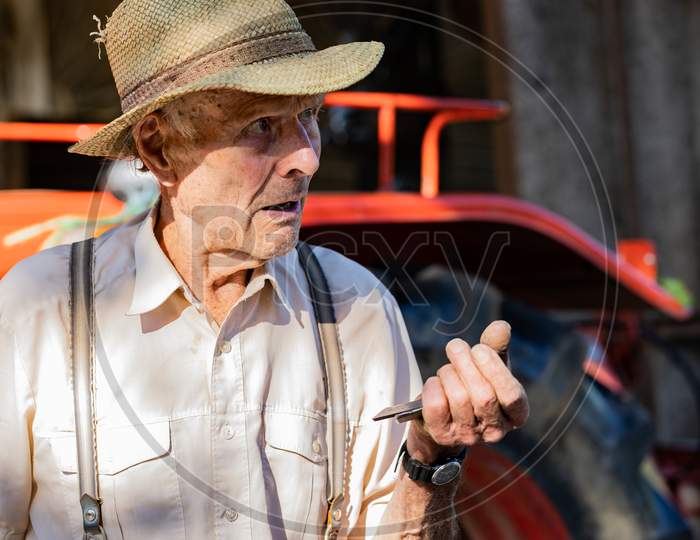 Portrait Of Very Old Farmer With Straw Hat Explaining Life In Front Of A Red Tractor.