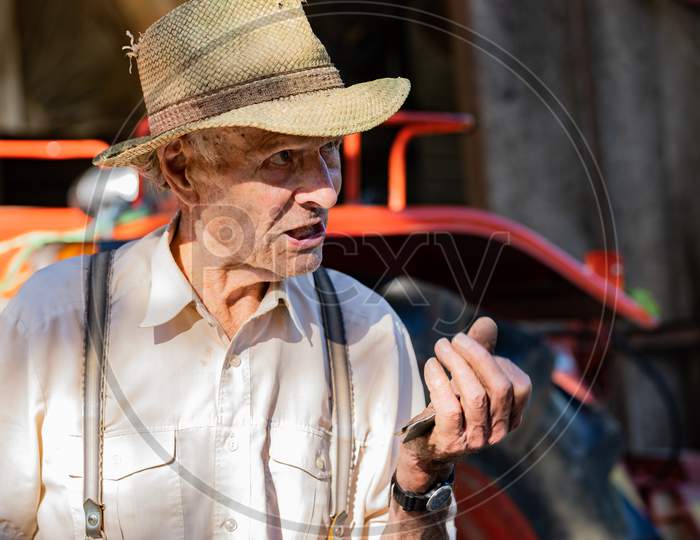 Portrait Of Very Old Farmer With Straw Hat Explaining Life In Front Of A Red Tractor.