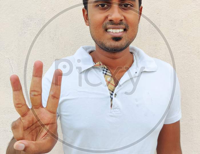 Smiling South Indian Young Man Wearing White Tshirt Pointing Up With Fingers Number Three. Isolated On White Background.