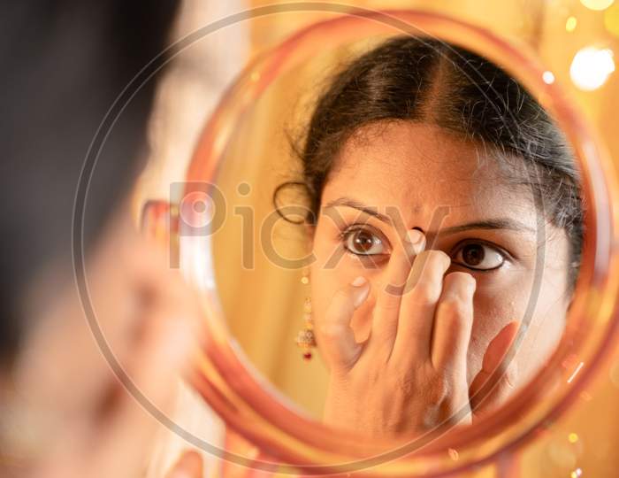 Indian Married Woman Applying Bindi, Sindoor Or Decorative Mark To Forehead In Front Of Mirror During Festival Celebrations With Decoration Lights As Background.