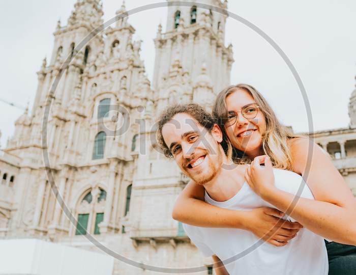 Young Couple Smiling To Camera With The Woman Over The Man In Front Of A Touristic Place