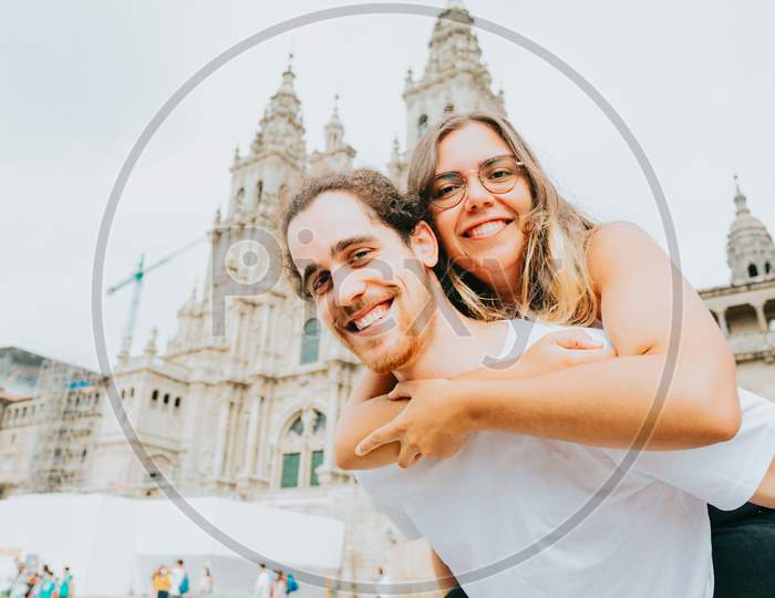 Young Couple Smiling To Camera With The Woman Over The Man In Front Of A Touristic Place
