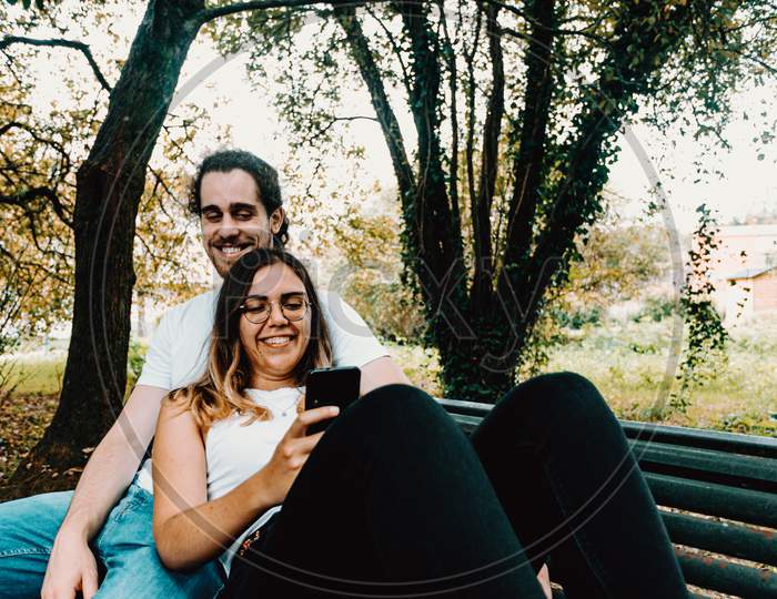 Young Couple Resting In A Bench While Looking The Phone In The Park