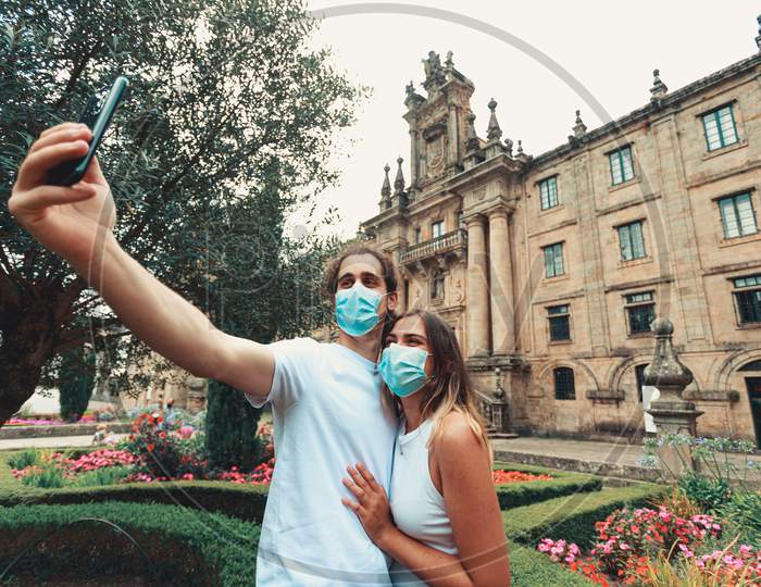 Young Couple Taking A Fancy Selfie With The Masks On In A Garden In Front Of An Old Building