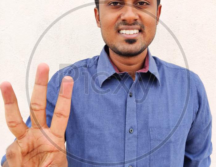 Smiling South Indian Young Man Wearing Blue Shirt Pointing Up With Fingers Number Three. Isolated On White Background.