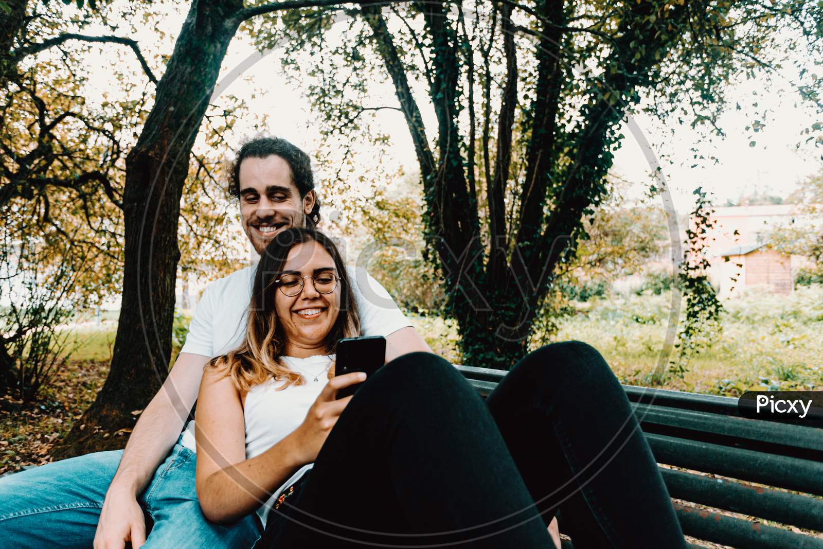 Young Couple Resting In A Bench While Looking The Phone In The Park