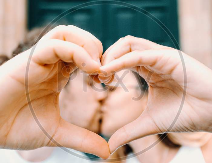 Young Couple Making A Heart Symbol With The Hand And Kissing Each Other On The Background