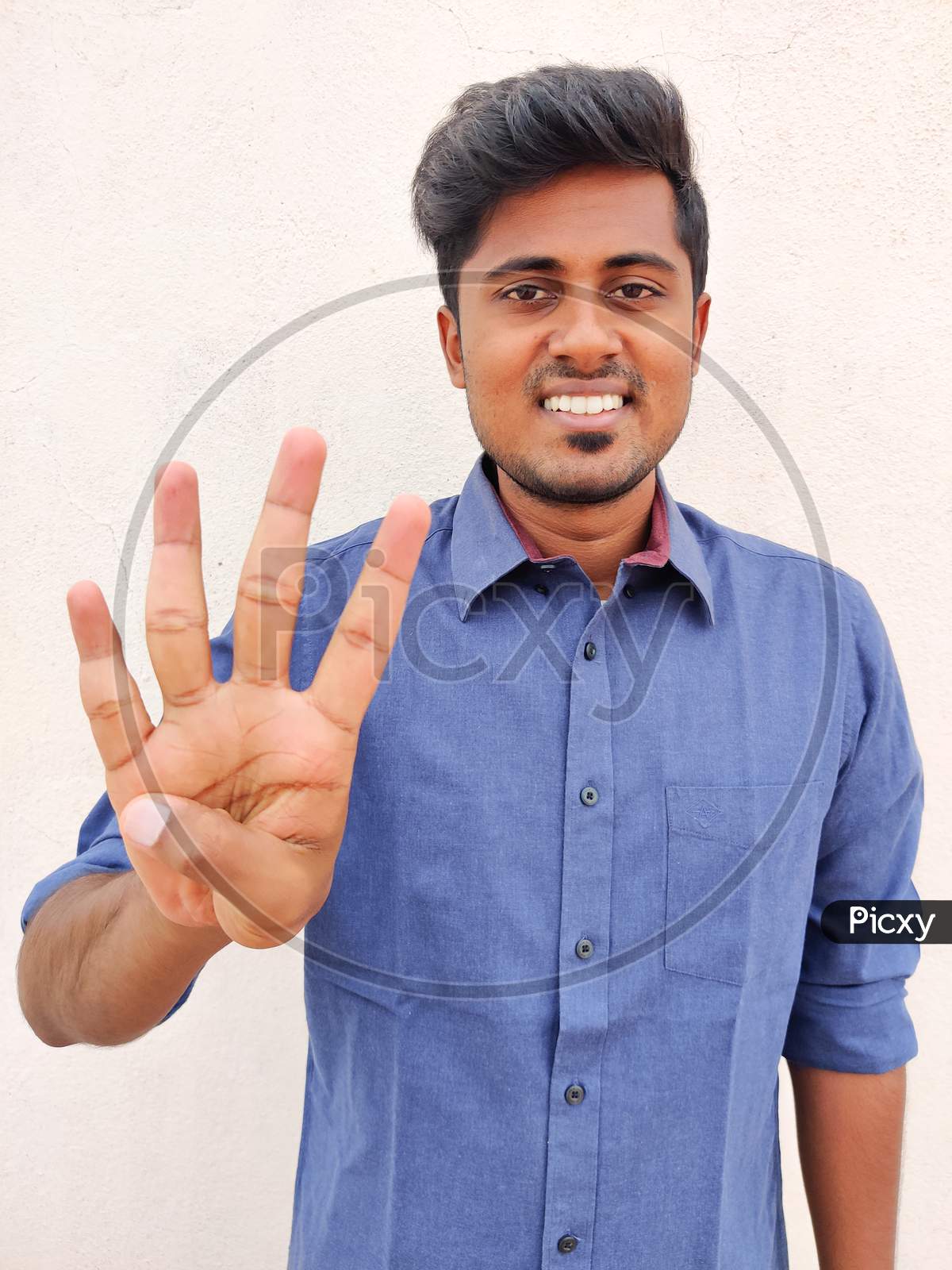 Smiling South Indian Young Man Wearing Blue Shirt Pointing Up With Fingers Number Four. Isolated On White Background.