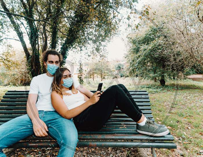 Young Couple With The Masks On Resting In A Bench While Looking The Phone In The Park