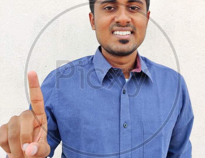 Smiling South Indian Young Man Wearing Blue Shirt Pointing Up With Fingers Number One. Isolated On White Background.
