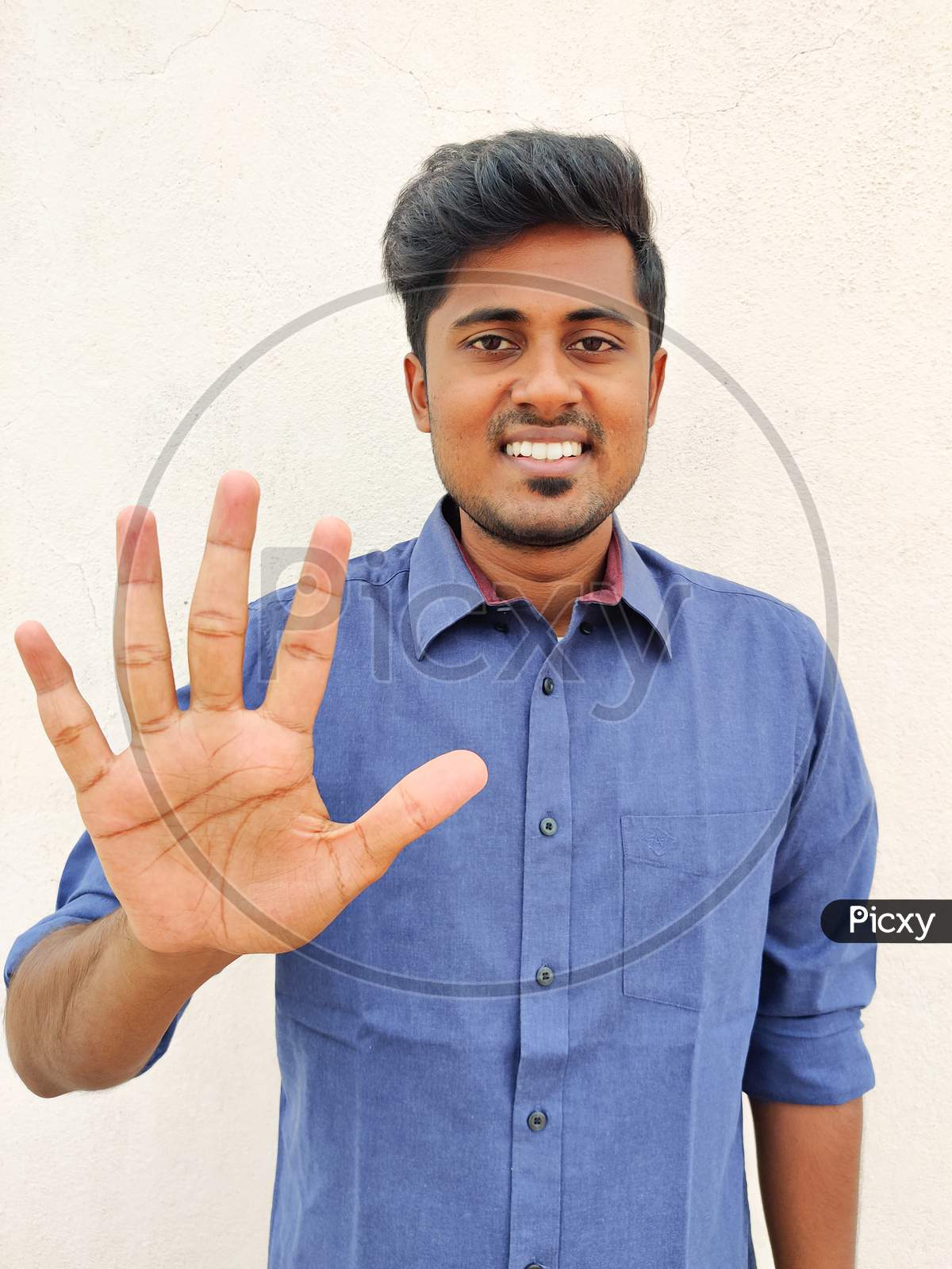 Smiling South Indian Young Man Wearing Blue Shirt Pointing Up With Fingers Number Five. Isolated On White Background.