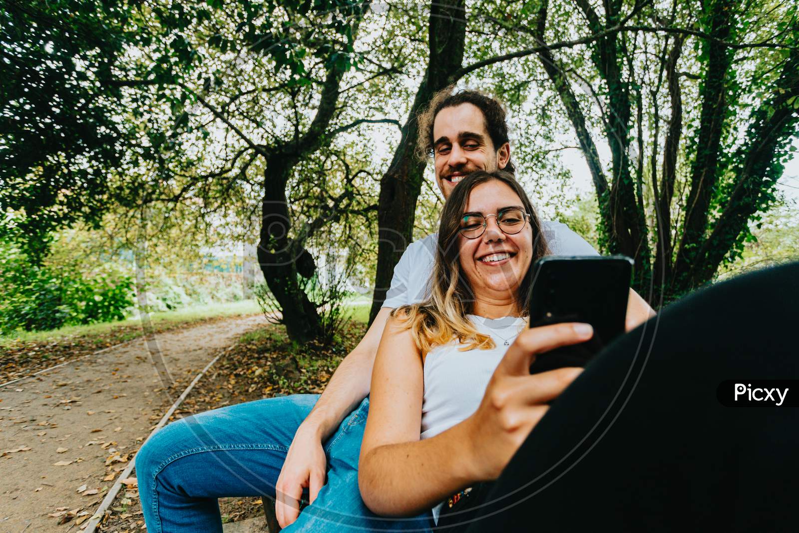Close Up Of A Young Couple Looking The Mobile Phone While Sitting On A Bench In The Park