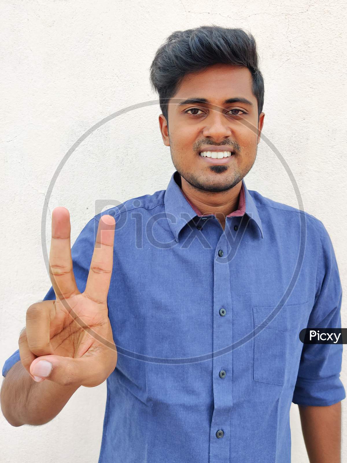 Smiling South Indian Young Man Wearing Blue Shirt Pointing Up With Fingers Number Two. Isolated On White Background.