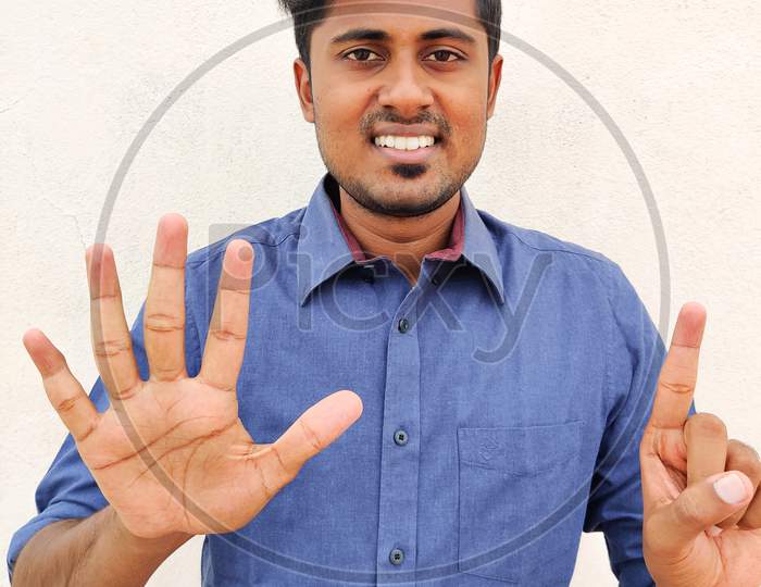 Smiling South Indian Young Man Wearing Blue Shirt Pointing Up With Fingers Number Six Or 51 Or 15. Isolated On White Background.