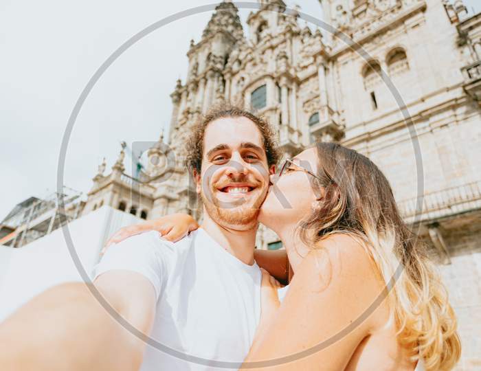 Horizontal Shot Of A Young Couple Kissing In Front Of A Cathedral During A Sunny Day