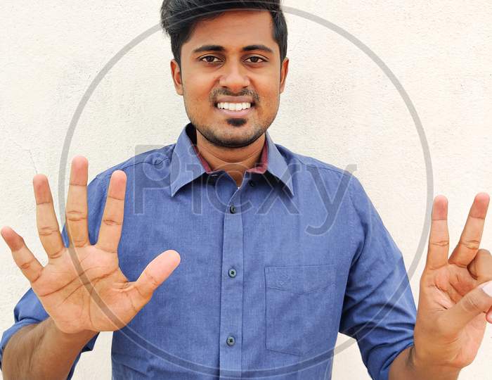 Smiling South Indian Young Man Wearing Blue Shirt Pointing Up With Fingers Number Seven. Isolated On White Background.