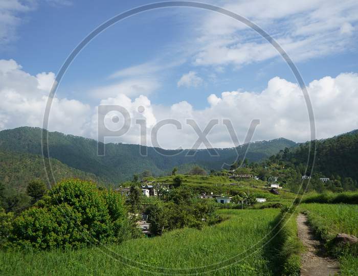 A Pathway In Between Green Fields With Blurred Mountains In The Background, Cloudy Blue Sky.