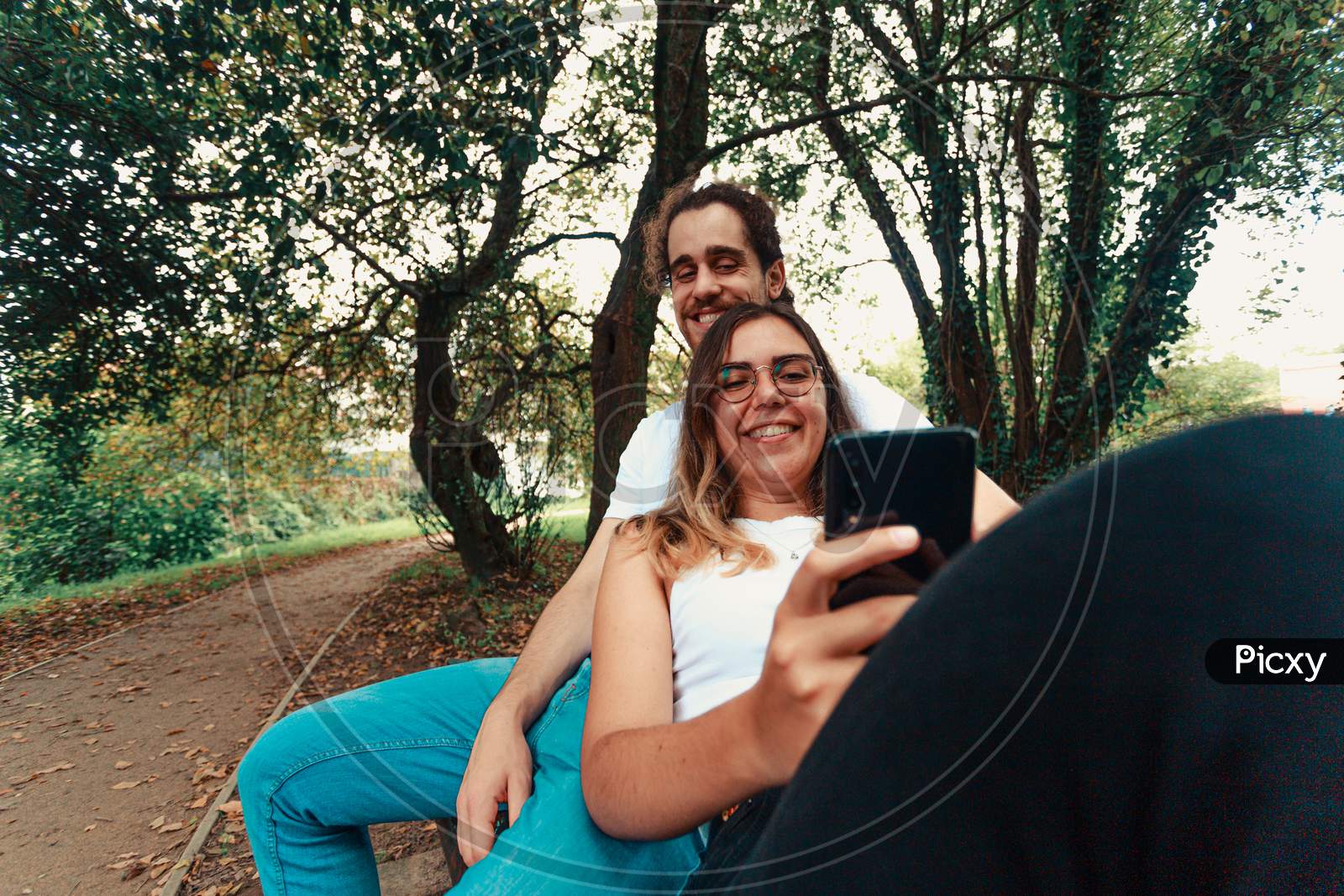 Young Couple Looking At The Smart Phone In A Bench On The Park While Smiling