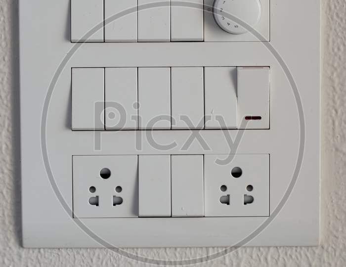 Designer Electrical White Switch Board With Regulator And Switches And Three Pin Sockets On The White Wall