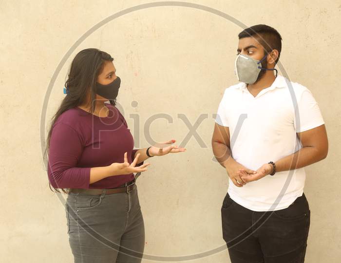 Young Indian Men And Women Discussing About Coronavirus (Covid 19) Topic While Both Wearing Protective Face Mask And Casual Clothes, Against Wall Background