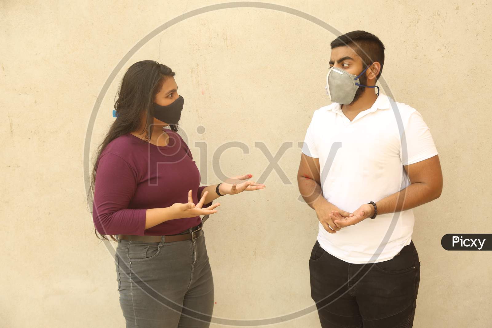 Young Indian Men And Women Discussing About Coronavirus (Covid 19) Topic While Both Wearing Protective Face Mask And Casual Clothes, Against Wall Background