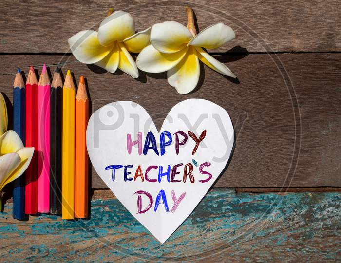 Happy Teacher'S Day Wish Written On Heart Shaped Paper Note With Color Pencils And Plumeria Flowers On Wooden Background, Perfect For Wallpaper