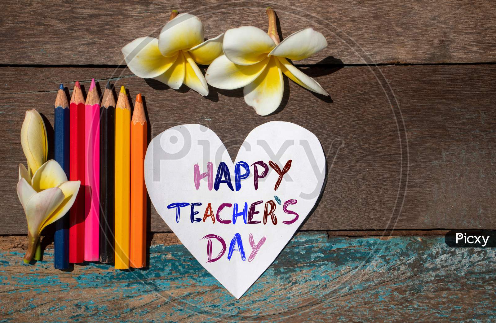 Happy Teacher'S Day Wish Written On Heart Shaped Paper Note With Color Pencils And Plumeria Flowers On Wooden Background, Perfect For Wallpaper