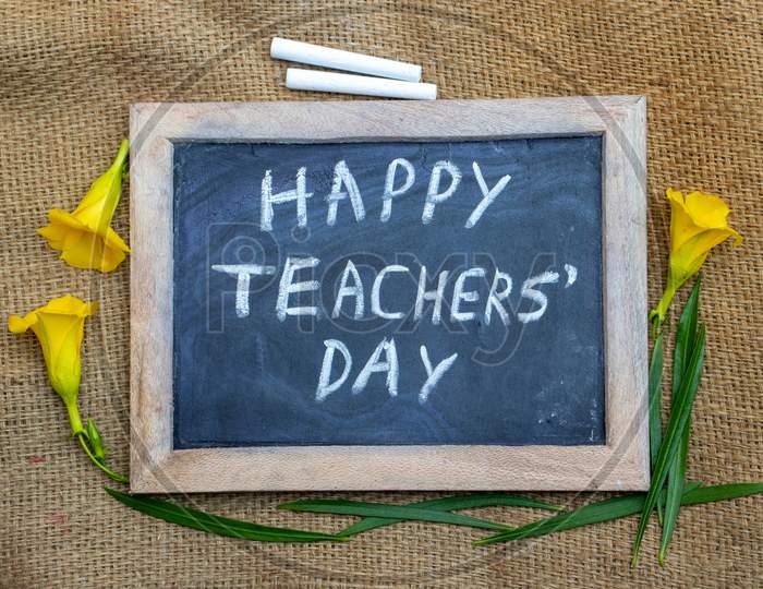 Happy Teachers' Day Written On Chalkboard With Yellow Oleander Flowers Isolated On Burlap Background, Perfect For Wallpaper