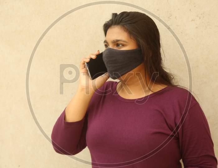 Happy Young Indian Beautiful Girl Wearing Protective Face Mask And Talking On Her Cell Phone, Standing Against Wall Background, Covid 19 Pandemic, Lock Down, Stay Home