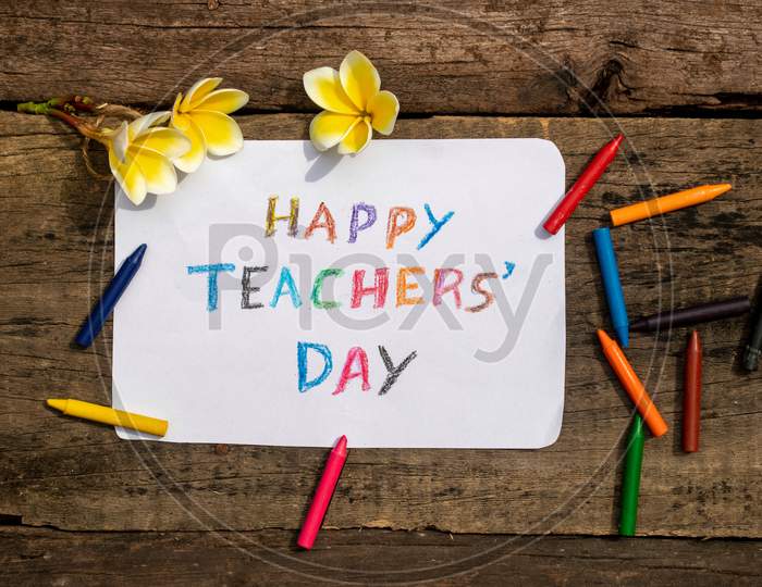 Happy Teachers' Day Wish Written On Paper Note With Color Pencil And Plumeria Flowers On Wooden Background