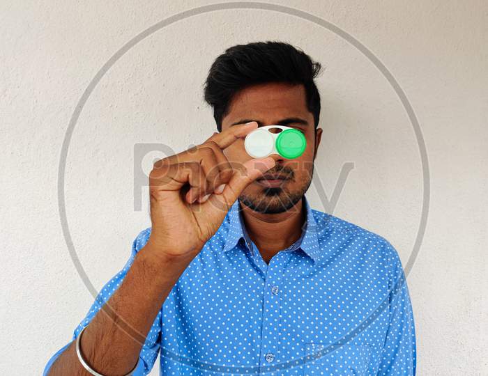 Handsome Man Dressed In Blue Shirt Holding Contact Lens Box In His Hands. Eye Wear.Daylight. White Background