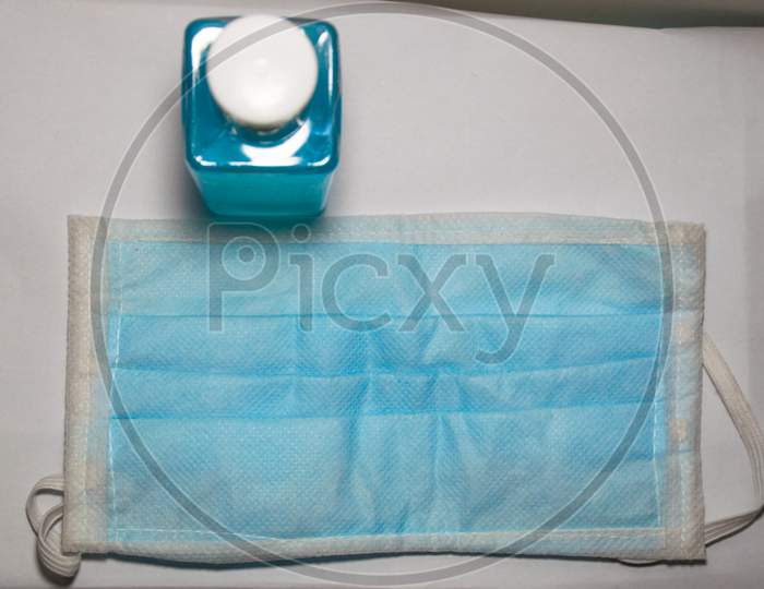Top View Of A Bottle Of Hand Sanitizer Gel And A Surgical Mask Required For Coronavirus Infection Prevention Placed On A White Table And A White Background