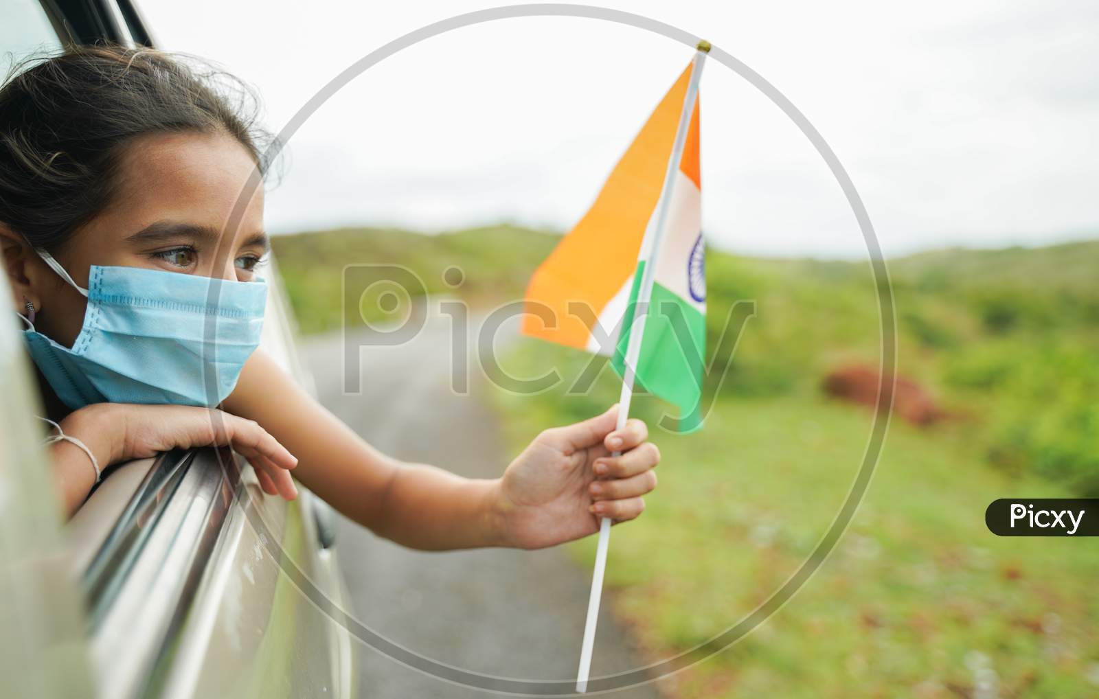 Young Girl Kid With Medical Mask Holding Indian Flag In Moving Car Window - Concept Of Celebrating Independence Or Republic Day During Coronavirus Or Covid-19 Pandemic