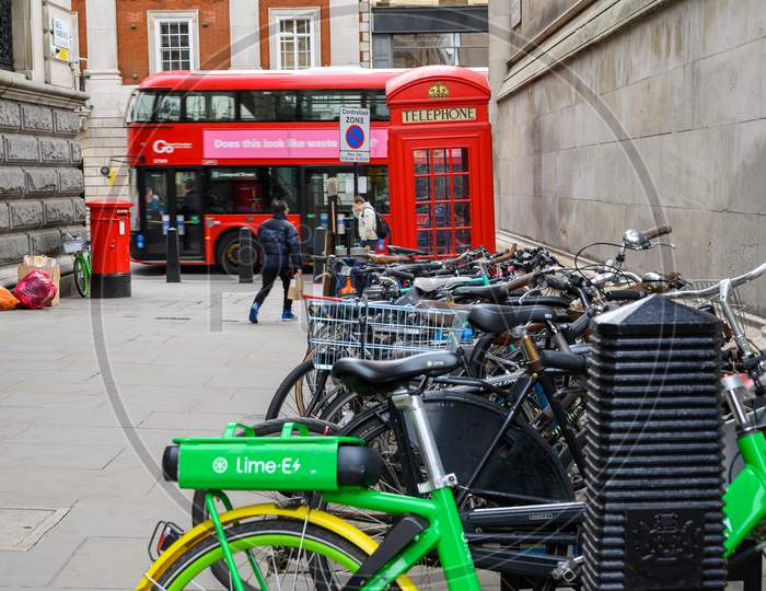 A Lime Electric Assist Rental Bike Parked With Regular Bicycles With A Red London Double Decker Bus, A Traditional Red Phone Box And Post Box In The Background