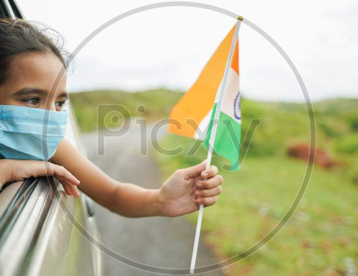 Young Girl Kid With Medical Mask Holding Indian Flag In Moving Car Window - Concept Of Celebrating Independence Or Republic Day During Coronavirus Or Covid-19 Pandemic