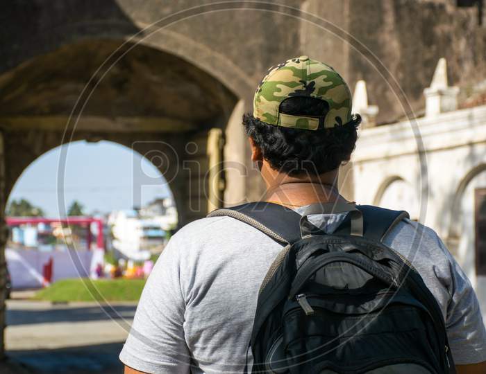Rear Portrait Of Young Indian Traveler With Backpack And Cap Looking At The Perimeter Wall Of A Fort In India