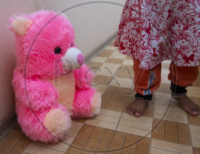 Close Shot Of Pink Teddy Bear Sitting On Floor With Baby Girl Legs Background