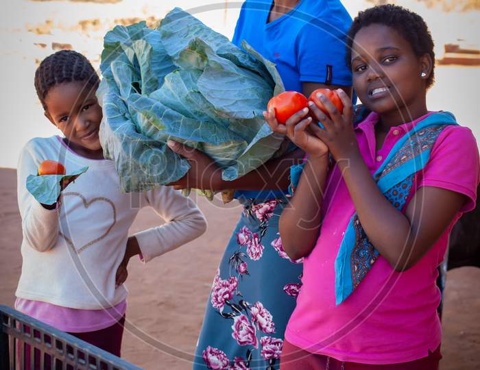 An African Woman With Children Holding A Cabbage And Tomatoes